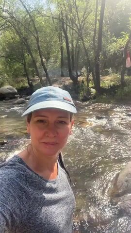 A woman with shoulder-length dark hair, wearing a light blue baseball cap and a gray t-shirt, smiles at the camera. She is standing by a shallow, clear stream with rocks and trees in the background. Sunlight filters through the trees, creating dappled light on the water. The water is flowing and appears to be in a natural setting. The woman's face is well-lit, and she has a relaxed expression. There are no other people visible in the frame. The overall impression is one of a serene outdoor environment.