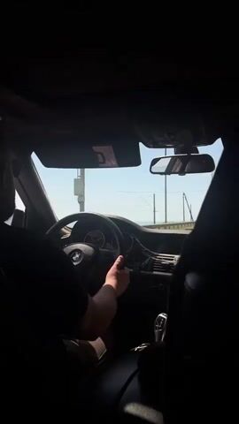 A person is driving a BMW car. The view is from the back seat, focusing on the driver's hands on the steering wheel. The interior of the car is mostly dark, contrasting with the bright, clear sky visible through the windshield. Power lines and trees are visible outside, indicating the car is moving, likely on a road.
