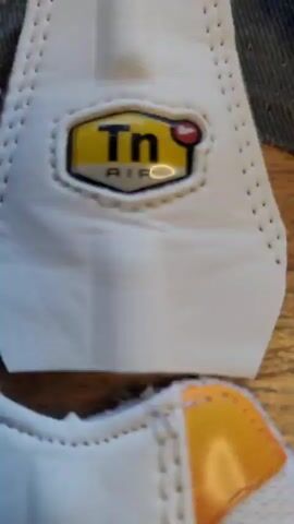 The video displays various disassembled components of what appear to be Nike Air Max Plus (Tn) sneakers, laid out on a wooden workbench. The pieces are primarily white leather, adorned with striking red and orange flame-like patterns, and some yellow accents. Several pieces prominently feature the recognizable "Tn Air" logo.

The camera pans across these individual sneaker parts, showcasing different sections of the shoe's upper, including the distinctive flame-shaped overlays, tongue sections, and heel components. The arrangement suggests a preparation stage for assembly or custom shoe creation. In the background, other materials like black fabric cut-outs and a mix of colorful stickers are visible on the wooden surface, along with glimpses of workshop tools such as a small vise or clamp. A faint Nike swoosh can be seen on one of the longer, side panel pieces. The overall scene indicates a detailed crafting or customization process related to footwear. The location context given is Sant Just Desvern, Spain.