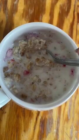 The video shows a close-up overhead view of a white mug filled with oatmeal. A spoon is stirring the oatmeal, creating ripples in the liquid. Small red pieces, possibly berries, are visible mixed in with the oatmeal. The mug is placed on a wooden surface, likely a table, with a visible wood grain texture. The lighting suggests an indoor environment.
