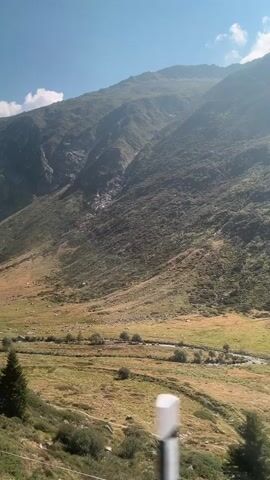 The video shows a mountainous landscape with a valley in the foreground. The mountain slopes are covered in greenery and rocks, with some visible signs of erosion or water flow. The valley is a mix of grassy fields and sparse vegetation, with a winding road or path visible. The sky is clear with a few wispy clouds. The camera appears to be moving, possibly from a vehicle, as evidenced by the slight blur and the changing perspective of the foreground elements. The lighting suggests it is daytime, likely morning or afternoon, with the sun casting shadows on the mountainside. There are no people or animals visible in the frame.