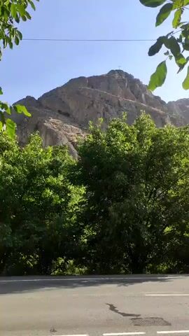 The video shows a tree with green leaves in the foreground, obscuring parts of the background. Behind the tree, a paved road is visible with white lane markings. A rocky mountain with a sheer face is in the distance, under a clear blue sky. There are also power lines and a metal tower present. A person is seen standing near the road, and further down the road, two people are walking away from the camera. The sun is shining brightly, casting shadows.