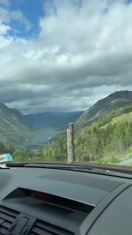 The video shows a view from inside a car, looking out through the windshield. The car appears to be on a high vantage point, overlooking a scenic valley. The valley has a lake or river running through it, surrounded by mountains and forested areas. The sky is partly cloudy with patches of blue. There is a wooden post visible in the foreground, near the side of the road. The dashboard of the car is visible, and it is a dark gray color. There are no visible people or significant actions occurring in the video.