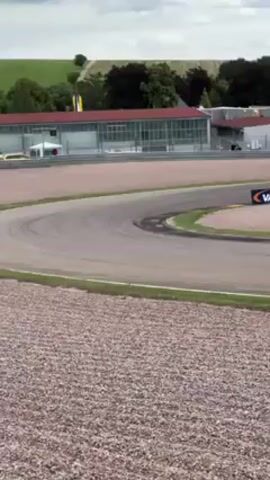 The video shows a black and yellow Porsche race car driving on a racetrack. The car is rounding a curve to the left, and the background includes buildings, a grassy hill, and a gravel area. The sky is overcast. A Vaico brand sign is visible.