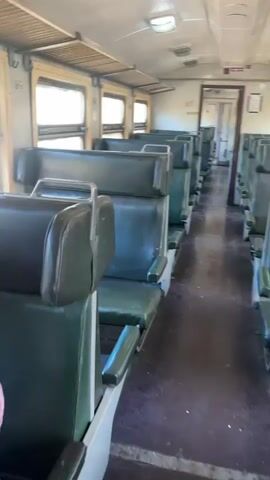 The interior of a train car is captured from the perspective of a person walking through it. The car features rows of green vinyl seats, arranged in pairs facing one another. Each seat has a small armrest. The windows are rectangular and allow natural light to filter into the car. The floor is dark, and the walls are a pale yellowish color. Overhead, there are lights recessed into the ceiling. At the far end of the car, there is an open doorway leading to another car. The overall aesthetic is dated, suggesting an older train. There are no people visible in the car. The camera moves slowly forward, showing more of the car's interior.
