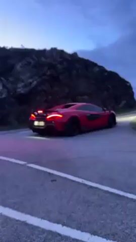 A red McLaren turns a corner on a rural road as a dark blue Porsche drives behind it. The road surface is asphalt with white lines, and the surroundings include a rocky hillside and dusk sky. The McLaren is shown with its brake lights on. The sound of the vehicles driving can be heard.