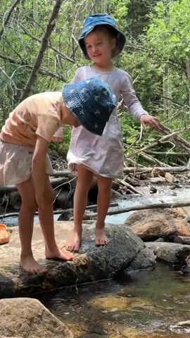 Two young girls, both wearing blue denim bucket hats with a bow pattern, are standing on rocks beside a stream. The girl in the foreground is wearing a peach-colored t-shirt dress and is bent over, reaching her hands into the water. The girl behind her is wearing a long-sleeved, light purple dress with a swirling pattern and is standing upright, looking towards the stream. Both girls are barefoot. The setting is a wooded area with trees and branches visible. A rocky stream with flowing water is in the background. The lighting suggests it is daytime, likely sunny. The overall atmosphere appears to be one of childhood exploration and enjoyment of nature. There is no visible text in the media.