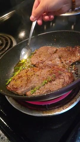 A person's hand, adorned with a beaded bracelet, is visible in the upper right corner of the frame. The hand holds a spoon, which is being used to baste two pieces of steak cooking in a black non-stick skillet. The steaks are seasoned with what appears to be pepper and herbs, and rosemary sprigs are also present in the pan. The skillet rests on an electric stovetop, which is turned on, indicated by a pink glow around one of the burners. The background is dark, suggesting a kitchen environment. The action focuses on the process of cooking the steak, with the spoon lifting and pouring oil over the meat.