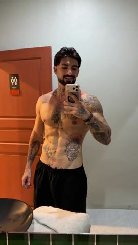 A shirtless man with tattoos is taking a selfie in a restroom. The man has dark hair and a beard and is wearing black shorts. He's holding a smartphone in his right hand, with his left hand resting casually on his hip. His abdominal muscles are visible and defined. He is smiling at his reflection in the mirror. The background includes a sink, a towel, and a red door with a restroom sign on it. The restroom is well-lit with a plain wall.
