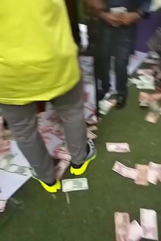 The video shows people standing on a grassy surface with scattered currency bills. Several individuals are wearing traditional West African attire in shades of maroon and yellow. One person wears a neon yellow shirt and grey pants. The men mostly stand, with some appearing to be dancing or moving to the music. The environment seems to be outdoors, possibly under a tent or awning.