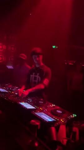 Two DJs are seen behind a DJ booth in a dimly lit club. Red lighting illuminates the scene. The DJ in the foreground, wearing a black t-shirt with a white graphic and the number 68, is actively mixing music on turntables and a mixer. He has tattoos on his arms. The second DJ, to his left and slightly behind, is wearing a cap and a dark shirt and appears to be observing or assisting. In the background, indistinct figures can be seen, and an "EXIT" sign is visible. The atmosphere is energetic and typical of a nightclub setting.