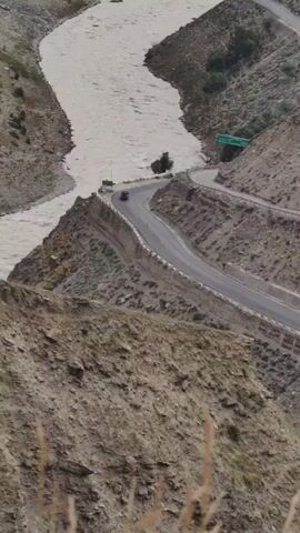 This video shows a winding road with a few cars driving on it, bordered by a wide, fast-flowing river. The road hugs the side of a steep, rocky mountain, with the river in the valley below. The landscape is rugged and arid, with sparse vegetation on the mountain slopes. In the distance, the river widens, and a small settlement is visible on its banks. There are also hints of buildings and structures along the river. The sky appears overcast, suggesting a cloudy day. The overall mood is one of natural beauty and a sense of isolation, with the road representing human presence within a vast, wild landscape. The location appears to be in a mountainous region, with the river carving its way through the valley. There is no visible text in the video.