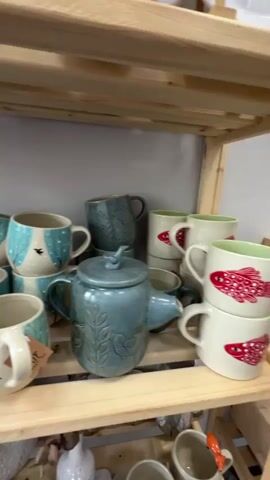 The video shows a close-up of shelves filled with handmade ceramic pottery. The items include mugs with blue and white tree designs, a teal teapot with leaf imprints, larger teal bowls, and stacks of white mugs with red designs. There are also smaller white ceramic pieces and a green ceramic gourd-like figure with a crown. The shelves appear to be made of light-colored wood, and the background is a plain gray wall. The lighting is soft and diffused, suggesting an indoor setting, likely a shop or studio. There are no people visible in the video.