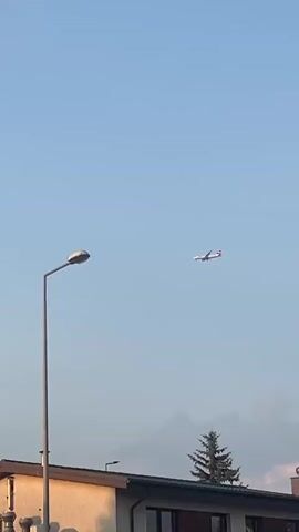 The media file is a vertical video showcasing a clear, pale blue sky on what appears to be a sunny day with a few scattered clouds. In the upper portion of the frame, a commercial airplane is visible, flying from left to right. In the lower section, a building with a light-colored facade and a brown-tiled roof is partially visible. A tall, grey lamppost stands prominently in the foreground, to the left of the frame. To the right of the building, a tall, dark green evergreen tree is silhouetted against the sky. The scene appears to be set in a suburban or semi-rural environment, possibly in Stara Iwiczna, Poland, given the provided context. The time of day seems to be late afternoon or early evening, indicated by the warm, soft lighting illuminating the side of the building and the underside of the clouds. There is no visible text in the media file. The overall mood is peaceful and serene.