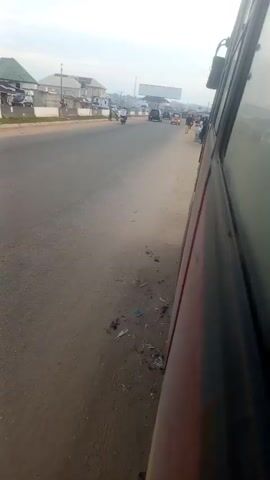 The video shows a view from inside a vehicle, looking out at a road and its surroundings. The perspective is from the right side of the vehicle, with the side of the vehicle, its red exterior, and tinted windows prominently featured. Outside the vehicle, the road stretches into the distance, with a dirt shoulder to the left. On the far side of the road, buildings and some trees are visible. Several people are walking along the side of the road, and a few vehicles, including a tricycle, are on the road. The sky appears overcast. The overall scene depicts a roadside environment, possibly in a town or city, with normal daily activity taking place. There is some litter on the dirt shoulder. The time stamp indicates that the video begins at 00:00 and ends at 00:04.