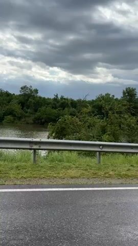 The video shows a view from a vehicle traveling along a wet road. On the left side of the road, there is a metal guardrail, followed by a grassy embankment and a body of water surrounded by dense green trees. The sky is overcast with dramatic, dark clouds. The vehicle's side mirror is partially visible in the bottom right corner of the frame, indicating forward motion. The water appears calm, and the overall environment suggests a natural, possibly rural setting under inclement weather.