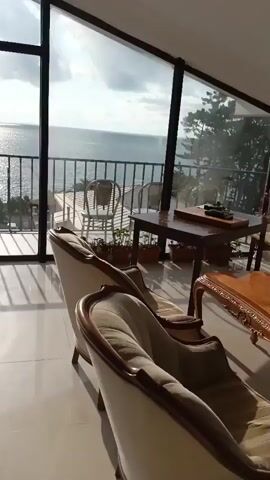 The video pans across a room with a large window overlooking the ocean. Inside, two ornate chairs are visible in the foreground. Through the window, a balcony with a railing, a chair, and some potted plants can be seen. Beyond the balcony, the ocean stretches to the horizon, with sunlight reflecting off the water. The sky is partly cloudy. Buildings and trees are visible on the land adjacent to the ocean.