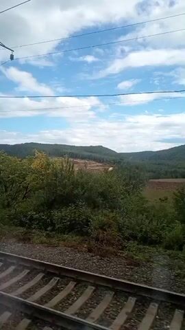 The video captures a view from a moving train, showing train tracks in the foreground. The landscape outside consists of rolling hills covered in dense green foliage and trees. The sky is blue with scattered white clouds. Overhead, power lines and their supporting poles are visible. In some areas, parts of the hills appear to have been cleared or disturbed, revealing patches of lighter-colored earth. The overall impression is of a journey through a natural, slightly rugged environment.