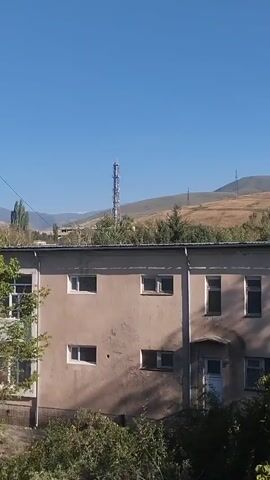 The video shows an exterior view of a pale, two-story building with multiple windows. The building appears to be old and weathered. In the background, there are rolling hills and mountains under a clear blue sky. A tall telecommunications tower is visible on one of the hills. The foreground is dominated by lush green trees and foliage, partially obscuring the lower portion of the building. The lighting suggests it is daytime, likely sunny. The camera is panning slowly from left to right, offering a panoramic view of the scene. There are no people visible in the video.