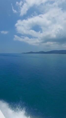 The video shows a view from a boat on the ocean. The water is a deep blue color and stretches out to a distant shoreline with green vegetation and rolling hills. The sky above is partially covered with large, billowing clouds that mix light and dark shades. White foam is generated in the lower left corner of the frame, suggesting the boat is in motion. The overall impression is one of tranquility and open space.