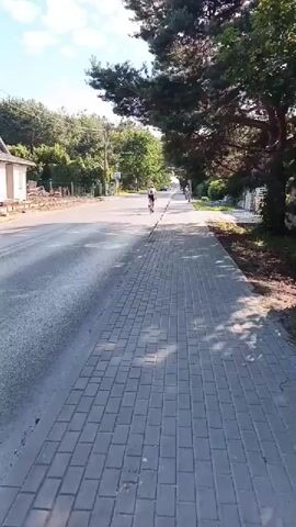 A person is riding a bicycle on a paved path next to a road. There is a woman walking on the path further ahead. The scene appears to be in a suburban or rural area with trees and houses visible. The sky is blue with some clouds.