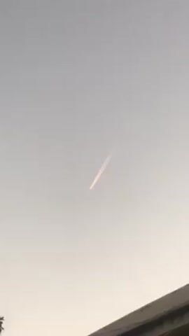 The video shows a light gray sky with a plane leaving a white contrail. The contrail is made up of two distinct lines, and stretches diagonally across the sky. The corner of a dark-colored building and a few branches of a tree are visible at the bottom of the frame. The overall lighting is dim.