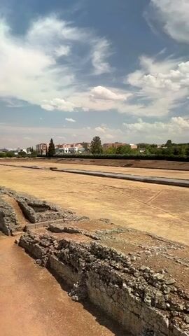 The video shows an aerial view of an archaeological excavation site on a sunny day with scattered clouds. In the foreground, there are ruins of stone walls and structures. The middle ground features a large, flat, excavated area with some demarcations and a small rectangular structure. In the background, there is a line of trees, followed by buildings and a large, tall, modern building resembling a silo or grain elevator. The overall impression is of an ancient site being preserved and studied amidst a modern urban landscape.