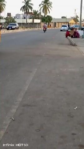 The video shows a street scene with moderate traffic. A yellow auto-rickshaw, followed by a motorcyclist wearing a red shirt with the number 9 on the back, travels down the road. Several parked cars are visible, and people are seated on the sidewalk. The road is grey and has a white line marking the lane. Palm trees line the road, and buildings are in the background. The video was recorded with an Infinix HOT 50i phone. The sky is light and overcast.