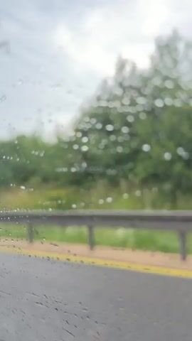The video shows a view from inside a vehicle looking out a window covered in raindrops. The car is on a road, indicated by the asphalt and yellow line. A metal guardrail runs along the side of the road, separating it from a grassy area and dense green trees beyond. In the distance, a large power pylon and electrical wires are visible against a cloudy sky. As the vehicle moves, the scenery passes by. At one point, a concrete structure with blue graffiti is visible under an overpass. The rain on the window distorts the view, creating numerous water droplets that streak down the glass.