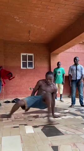 A young, shirtless man, wearing blue denim shorts and barefoot, performs a dynamic series of acrobatic and breakdancing moves on a tiled floor. He executes handstands, leg sweeps, and various ground-level movements with agility and energy.

Three other men are present in the scene, observing the performer. To the left, a man in blue jeans is seen putting on or taking off a red long-sleeved shirt. In the background, a man wearing a green t-shirt and light-colored shorts stands attentively, watching the performance. Further to his right, another man in a light-colored collared shirt and blue jeans also observes.

The setting appears to be an indoor or covered outdoor area, characterized by red brick walls and a matching brick-colored ceiling. A white-framed window is visible on the back wall, suggesting natural light is entering the space. The floor is a mosaic of irregularly shaped, multi-colored tiles, some of which appear worn or damaged. A single, bare light fixture hangs from the ceiling. A black cap and a small black bag rest on the floor to the left. The scene is brightly lit by natural daylight, indicating it is daytime. The location is Kuru, Nigeria.