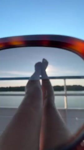 The video begins with a close-up of sunglasses with tortoiseshell frames held up against a clear blue sky. The camera then pulls back to reveal a pair of bare legs and feet propped up on a railing. Beyond the legs, a calm body of water is visible, with a tree-lined horizon in the distance. The sun is shining brightly, creating a glare on the water and casting rays through the lens of the sunglasses in the first frame. The overall impression is one of relaxation and enjoying a sunny day outdoors, possibly on a boat or ferry.