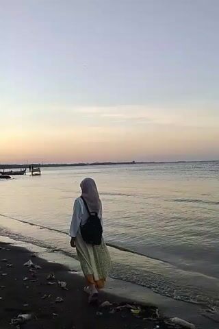 The video shows a person from behind, walking along a beach towards the right of the frame. The person is wearing a hijab, a light-colored long-sleeved shirt, and a patterned skirt. They also have a backpack on. The beach is dark and appears to have some debris on it. The ocean is to the right of the person, with gentle waves rolling in. The sky is a pale orange and pink, suggesting it is either sunrise or sunset. In the distance, on the left side of the frame, there are a couple of small boats docked. The overall mood is peaceful and serene.