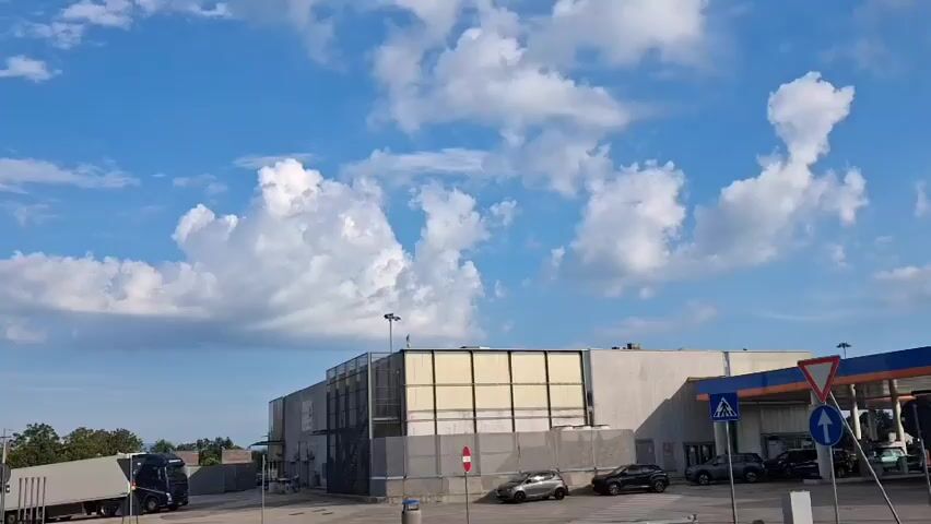 The video pans across a landscape under a bright blue sky with fluffy white clouds. In the foreground, a building with a grey facade and a white roof is visible, along with several parked cars. To the left, a large truck is parked. The background features rolling hills and mountains, with some trees and smaller buildings scattered throughout. A cell tower is also visible on the left side of the frame. The scene appears to be a mix of industrial and residential areas, with natural landscapes in the distance.