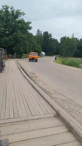 An orange truck is driving down a paved road bordered by a wooden walkway on the left and trees and grass on the right. A person is visible on the right side of the road in the distance. The sky is cloudy.
