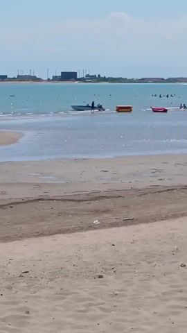 The video shows a beach scene with shallow water. In the foreground, there is sand with some debris. In the middle ground, shallow water covers the sand, with some puddles visible. Further out, there are several objects in the water: a small blue boat with a person in it, a yellow float, and a red float. In the background, there is a larger red boat with multiple people on board and a few people swimming near it. The sea is a turquoise color, and the sky is clear and blue. On the far horizon, there is a landmass with some buildings.