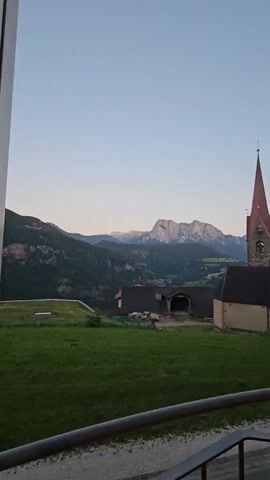 Here is a concise description of the video:

The video presents a static, wide shot of a picturesque landscape, likely taken in the European countryside. The foreground features a metal railing, suggesting the view is from a balcony or elevated platform. Beyond the railing is a lush green field, leading to a cluster of buildings that appear to be farmhouses or rural dwellings. Prominently visible is a church with a distinctive red-pointed steeple.

In the background, imposing mountains are partially obscured by clouds or mist. The sky above is a pale blue, indicative of early morning or late evening light. The overall ambiance is tranquil and scenic.