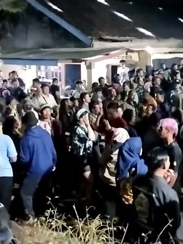 A crowd of people are gathered outdoors at night. Many of the individuals are looking upwards, towards something out of frame. There is a structure behind them with a corrugated metal roof. Some people are wearing headscarves.