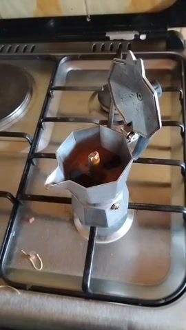 The video shows a moka pot brewing coffee on a stovetop. The pot is silver and the lid is open. The coffee is dark brown and frothy, with small bubbles visible. The stovetop is silver with black grates. There is a small hook laying near the pot. The camera zooms in slightly.