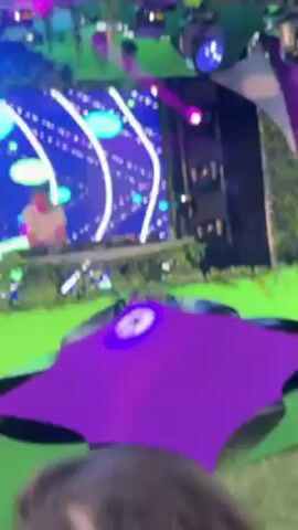 A man in a white t-shirt is DJing on a stage with a large LED screen behind him. The screen displays colorful abstract graphics.  The stage is decorated with greenery, and there are large purple shade canopies in front of the stage.  There are flashing lights above the stage.