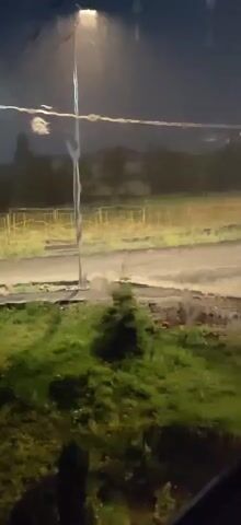 The video shows a streetlamp in the dark, with rain streaks visible. A small evergreen tree is in the foreground, and a dirt road is behind it. There are yellow fences in the background, and some buildings can be seen through the trees. A person is briefly visible walking along the road in the distance.