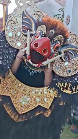 The video shows a close-up of a person wearing a large, elaborate mask that resembles a mythical creature or a demon. The mask is intricately designed with vibrant colors, gold detailing, and what appears to be real animal fur. The creature has a red snout with large nostrils, sharp tusks, and wide, expressive eyes. Decorative elements adorn the mask, including what look like ornate ear coverings and a headdress. The person inside the mask is wearing a black t-shirt. The background is slightly blurred but suggests an outdoor setting, possibly a street or courtyard, with a building and some greenery visible. The lighting is bright, indicating it is daytime. The person inside the mask moves their head, causing the mask to shift slightly, and their hands are visible near the sides of the mask.