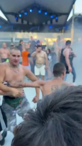 A group of people are gathered at an outdoor event, possibly a party or festival. The foreground is occupied by the backs of several people, their faces obscured. In the midground, a shirtless man with short hair and tattoos on his arm is dancing. He is wearing camouflage shorts. To his left, another shirtless man with tattoos on his back is being touched on the shoulder by a person whose face is not visible. Further back, a crowd of people are dancing and interacting. Some are shirtless, while others wear casual clothing. The environment appears to be an open-air venue with a stage or elevated platform in the background, equipped with lighting. Smoke or fog effects are present, creating a hazy atmosphere. The overall impression is that of a lively gathering with music and dancing.