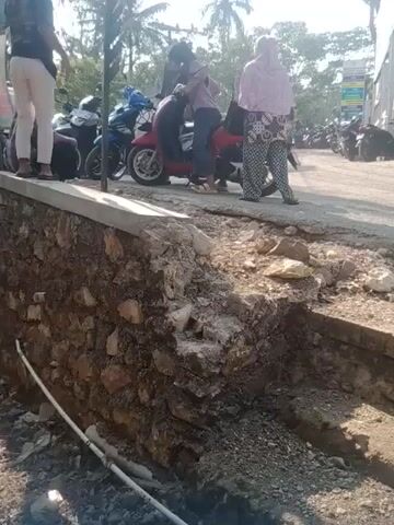 The video shows a partially collapsed embankment on the right side of the frame, revealing exposed rocks and soil. A concrete curb runs along the top of the embankment. On the left side of the frame, several motorcycles are parked on a paved area that appears to be a street or parking lot. Several people, some wearing headscarves, are standing near or sitting on the motorcycles. The scene is outdoors, with sunlight casting shadows, suggesting it is daytime. Vegetation, including palm trees and some leafy plants, is visible in the background on the left. The overall impression is of a public area with some damage to the infrastructure.