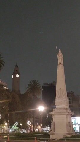 Here is a description of the video. 
The video shows a statue on top of a tall column, set against a night cityscape. The statue depicts a person holding a long object. To the left, a clock tower is visible, and a palm tree adds to the urban landscape. Streetlights illuminate the area, and other buildings can be seen in the background. A person wearing a uniform and a cone on the road suggests some level of activity or maintenance in the area. The sky is dark, giving the scene a nighttime ambiance.