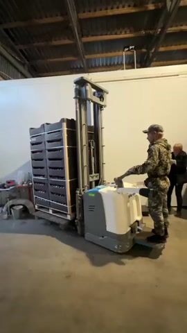 A man wearing camouflage clothing and a cap operates a powered pallet jack inside a warehouse. The pallet jack is lifting a tall stack of black crates filled with red fruit, likely apples. The crates are secured with straps to a wooden pallet. The warehouse has a concrete floor, metal ceiling beams, and white walls. Another person is partially visible in the background, standing near a wall. The pallet jack has a white and gray body with blue accents and a control handle. The man appears to be moving the stack of crates forward and upward.
