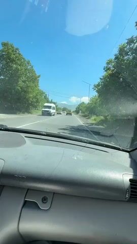 The video shows a view from inside a car traveling on a road on a sunny day. The camera is positioned behind the dashboard, looking out through the windshield. Several cars are visible on the road ahead, including a white van and two yellow cars. The road is lined with green trees and vegetation, and there are power lines overhead. In the distance, mountains can be seen under a clear blue sky. The car appears to be moving at a moderate speed.