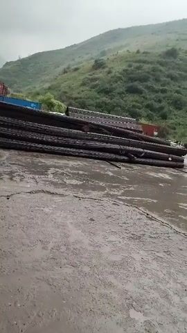 The video shows an outdoor scene with a concrete ground that appears wet or muddy. In the foreground, a portion of a black object, possibly a vehicle or equipment, is visible on the left side. In the midground, a large stack of dark, long, cylindrical objects, resembling rebar or logs, is laid out horizontally. Behind the stack, several shipping containers, one blue and others less distinct, are visible. The background features a steep, green, vegetated hillside under an overcast sky, suggesting a mountainous or hilly terrain. The overall impression is of an industrial or storage area in a natural, possibly remote, environment, possibly during or after rainfall.