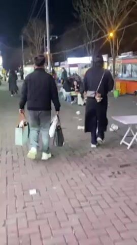 The video shows an outdoor night scene. Two people walk away from the camera on a brick sidewalk. The person on the left carries a light-colored tote bag and a black bag; they wear a black jacket, gray pants, and white shoes. The person on the right wears a black overcoat and carries a shoulder bag. There is a lot of trash strewn on the sidewalk. Several people are sitting at small tables with food stalls in the background. Streetlights are visible.