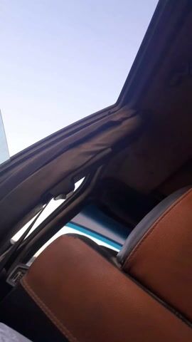 From a low vantage point, the camera is positioned inside a car. The interior features brown leather-like seats with black accents, and the car's frame is visible. The roof appears to be open, revealing a clear, light blue sky. There is ambient noise in the background.