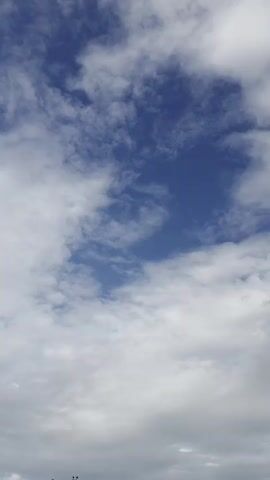 The video displays a time-lapse recording of the sky. The sun is not visible. Clouds are present, ranging from white and fluffy to grey and overcast. Patches of blue sky are visible between the clouds. The camera is positioned looking upwards, capturing the movement of the clouds across the sky. In the very bottom corners of the frame, trees and a lamppost are visible. The video appears to be recorded in an outdoor setting.